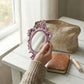 A hand holding a dusty pink ornate mirror near a linen makeup bag and leather journal on a white wooden table.