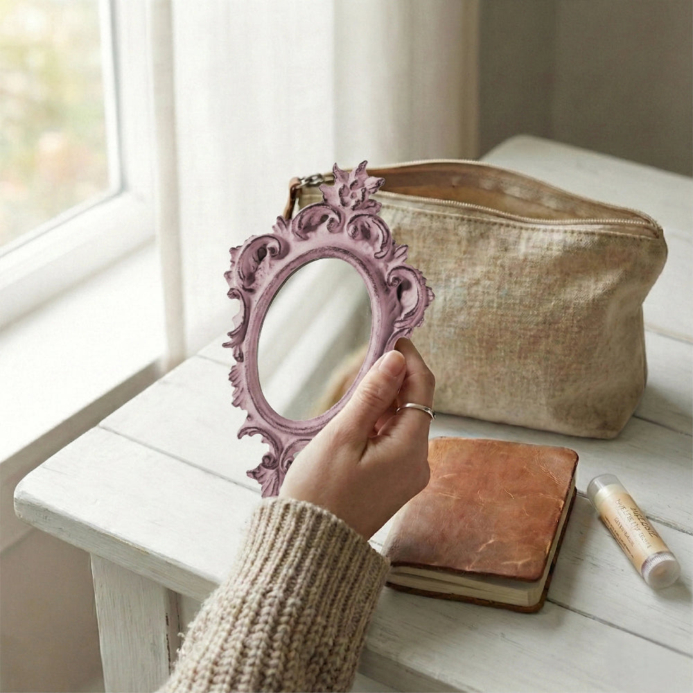 A hand holding a dusty pink ornate mirror near a linen makeup bag and leather journal on a white wooden table.