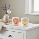 Two heart-shaped candle holders with lit candles on a white surface, next to a vase with flowers.