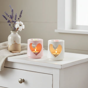 Two heart-shaped candle holders with lit candles on a white surface, next to a vase with flowers.