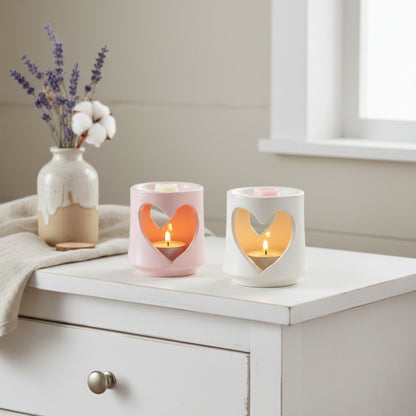 Two heart-shaped candle holders with lit candles on a white surface, next to a vase with flowers.