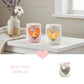 Two heart-shaped candle holders with lit candles on a white surface, with a vase of flowers in the background.