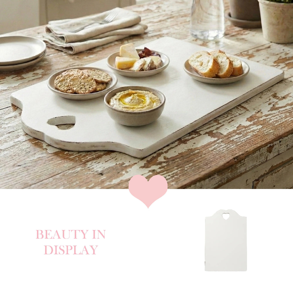 White rectangular serving board with a heart-shaped handle cutout featuring small bowls of hummus, cheese, crackers, and bread on a rustic distressed wooden table.