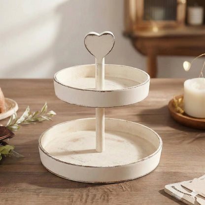 Two-tier antique white wooden tray with a heart-shaped handle on a wooden table with a candle and greenery.