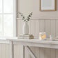 White heart-cutout wax melter with a lit candle, a bird figurine, and a vase with eucalyptus branches on books over a wooden table.