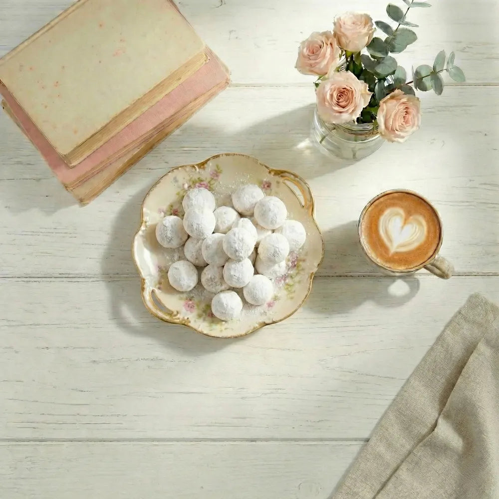 Assorted kahk on a plate with a cup of coffee, pink roses, and a book on a light wooden surface.