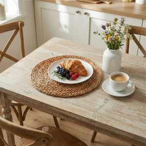 Natural halfa grass round placemat under a white ceramic plate and linen napkin on a white distressed wooden table with a farmhouse-style window background.