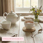 Natural round woven coaster under a white mug on a distressed light wood table with a white teapot and a jar of wildflowers.