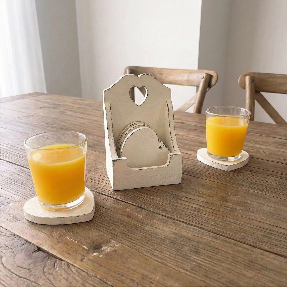 Two glasses of orange juice on heart coasters next to a tall white wooden holder on a natural wood table.