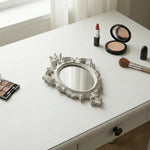 Ornate antique white oval mirror with distressed edges on a white vanity table next to makeup brushes and powders.