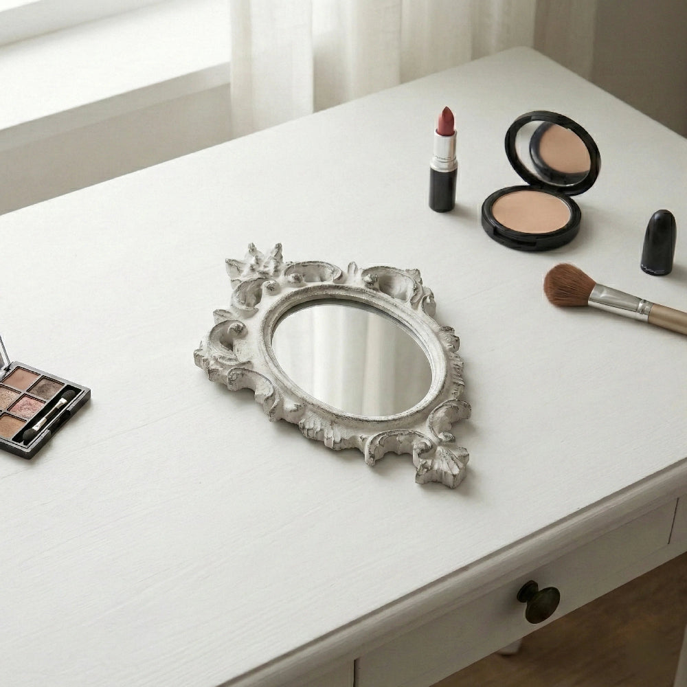 Ornate antique white oval mirror with distressed edges on a white vanity table next to makeup brushes and powders.