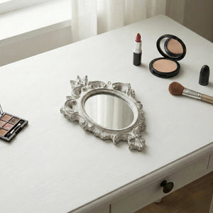 Ornate antique white oval mirror with distressed edges on a white vanity table next to makeup brushes and powders.