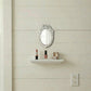 Ornate white oval mirror above a shelf with lipstick, nail polish, and a ring box on a cream shiplap wall.