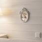 Small ornate oval mirror on a white shiplap wall reflecting pink pillows and a bed.