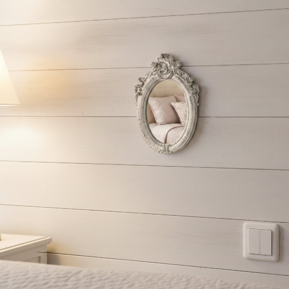 Small ornate oval mirror on a white shiplap wall reflecting pink pillows and a bed.