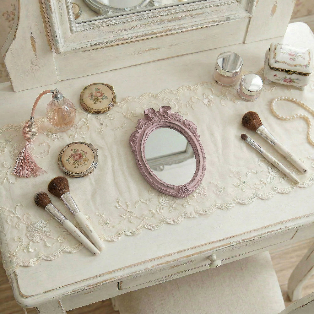 Ornate pink mirror on a lace runner atop a distressed white vanity with makeup brushes, perfume, and jewelry boxes.