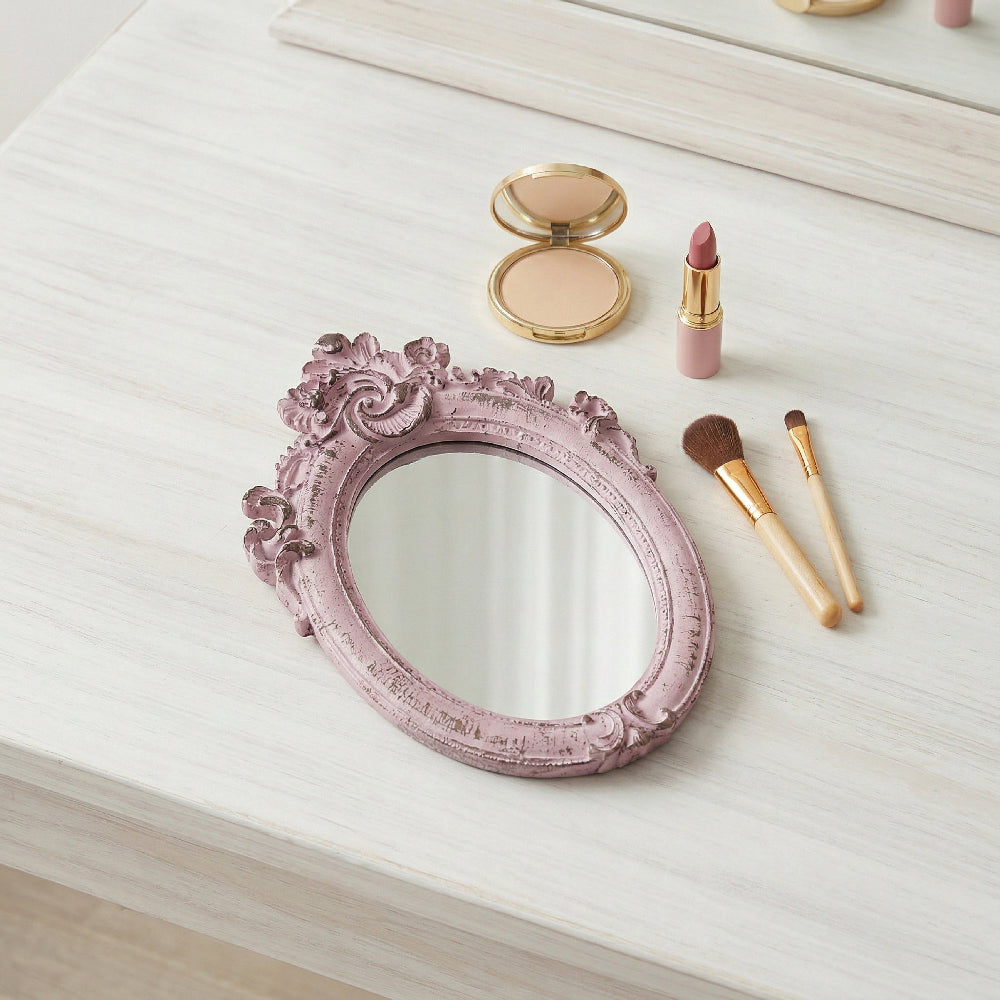Ornate pink oval mirror on a white wood vanity next to makeup brushes, lipstick, and a gold compact.