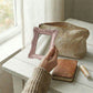 Hand holding a pink ornate rectangular mirror over a white wood table with a makeup bag and leather journal.