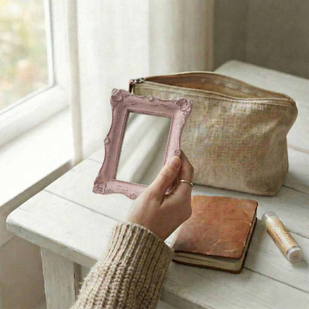 Hand holding a pink ornate rectangular mirror over a white wood table with a makeup bag and leather journal.