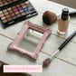 Small ornate pink rectangular mirror on a white wood table with an eyeshadow palette, makeup brushes, and foundation.