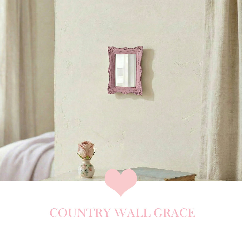 Small ornate pink rectangular mirror on a beige wall above a table with a pink rose in a vase and a book.