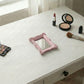 Ornate pink rectangular mirror on a white vanity desk alongside an eyeshadow palette, lipstick, and a makeup brush.
