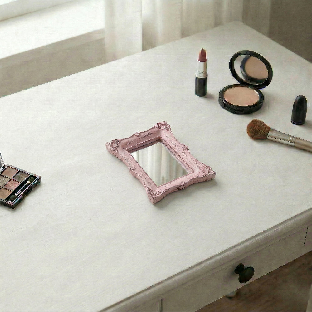 Ornate pink rectangular mirror on a white vanity desk alongside an eyeshadow palette, lipstick, and a makeup brush.