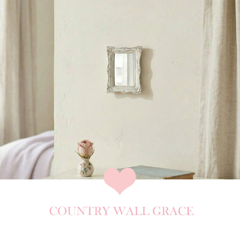Small ornate rectangular mirror on a beige wall above a table with a pink rose in a vase and a book.