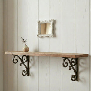Small ornate white square mirror mounted on a white shiplap wall above a wooden floating shelf with decorative iron brackets.