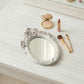 Ornate white oval mirror on a light wood surface with a compact, pink lipstick, and two makeup brushes.