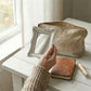 Hand holding an ornate white rectangular mirror over a wooden table with a makeup bag, leather journal, and lip balm.