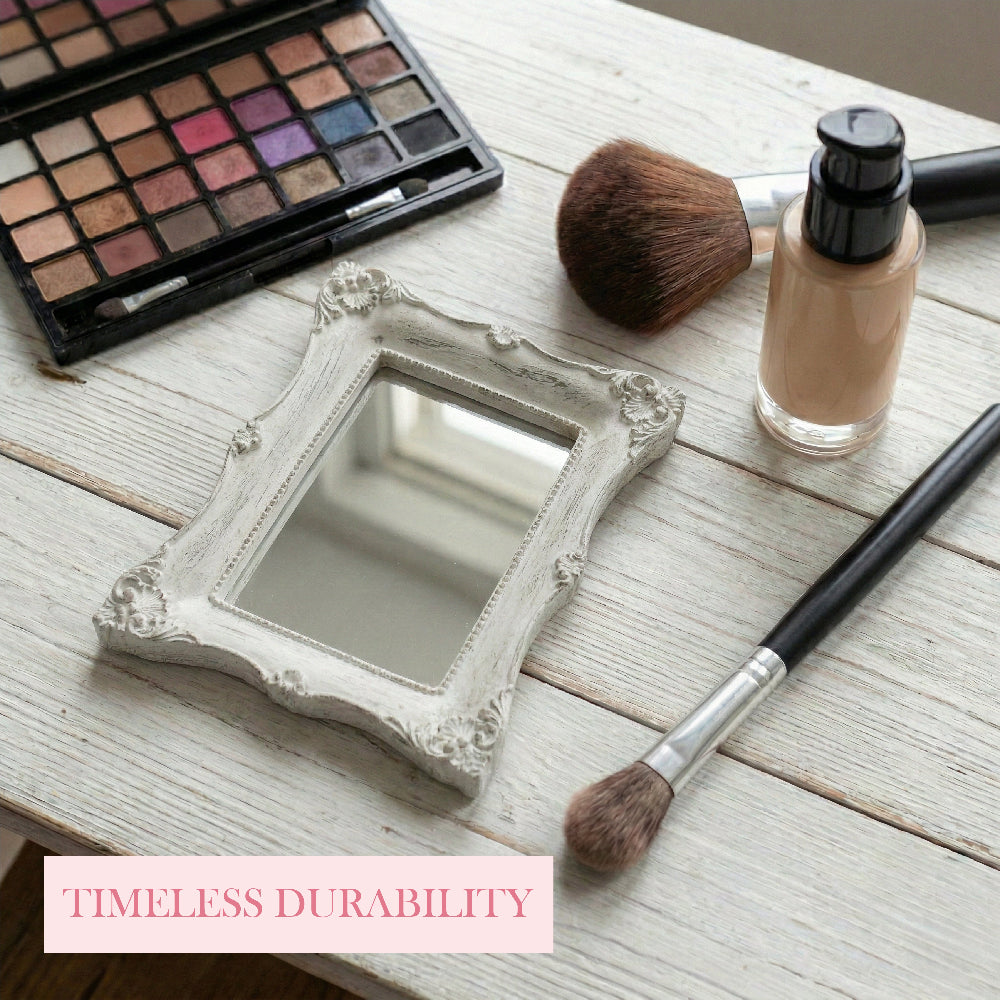 Small ornate rectangular mirror on a white wood table with an eyeshadow palette, makeup brushes, and foundation bottle.