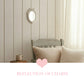 Small oval antique white mirror on a cream paneled wall above a distressed wooden chair with beige pillows.