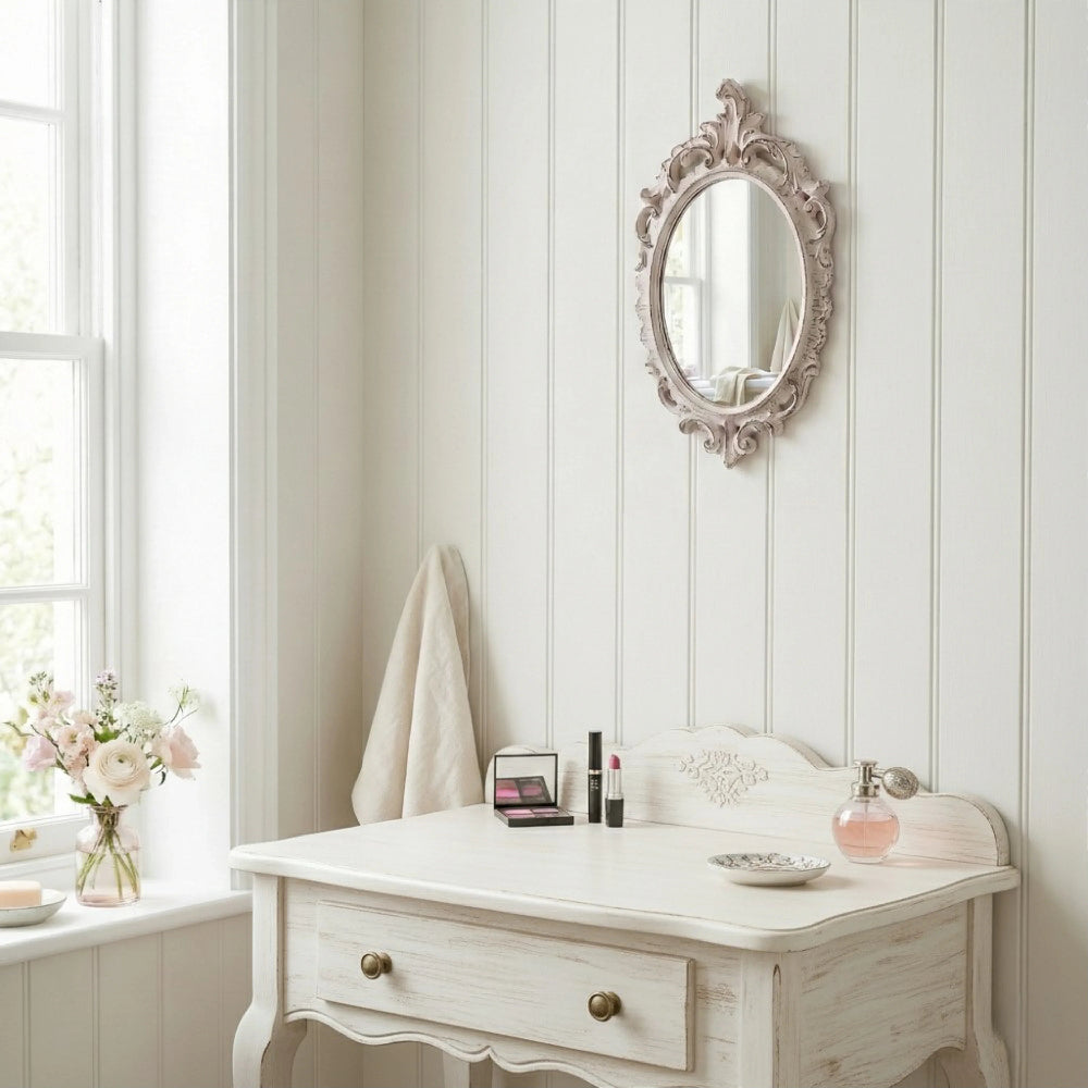 Oval antique white ornate mirror hanging on a vertical wood plank wall above a white vanity with makeup and perfume.