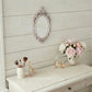 Oval antique white ornate mirror on a white wood wall above a vanity with peonies, makeup brushes, and perfume bottles.
