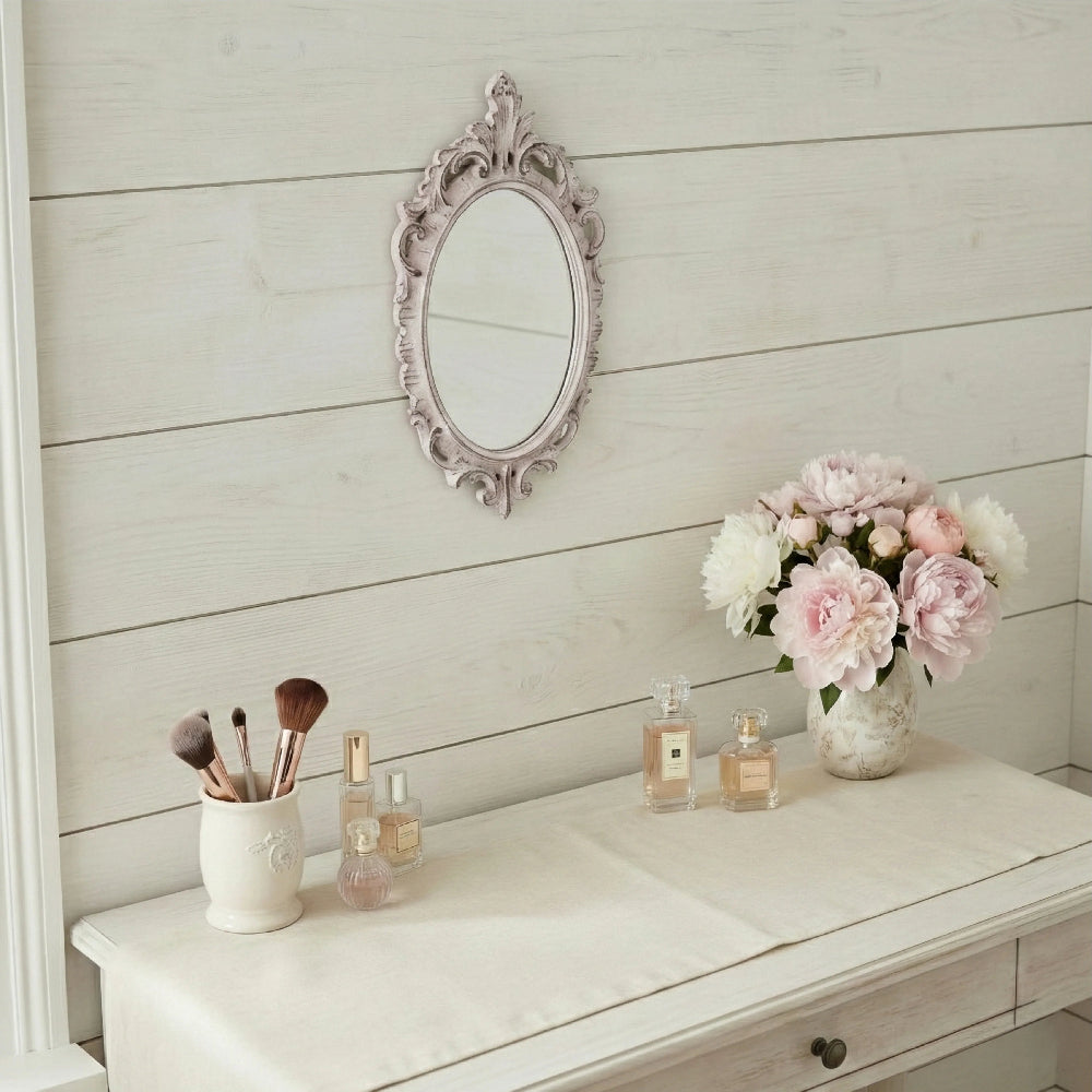 Oval antique white ornate mirror on a white wood wall above a vanity with peonies, makeup brushes, and perfume bottles.