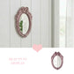 Oval pink ornate mirror with a ribbon top on a white paneled wall next to a window and text that says Beauty in Display.
