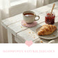 Pale pink woven coaster with a tea mug, croissant, and jam jar on a distressed white wooden table by a window.