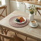 Pale pink halfa grass round placemat under a white plate with a croissant and berries, set on a distressed wood table with a coffee cup and wildflower vase.