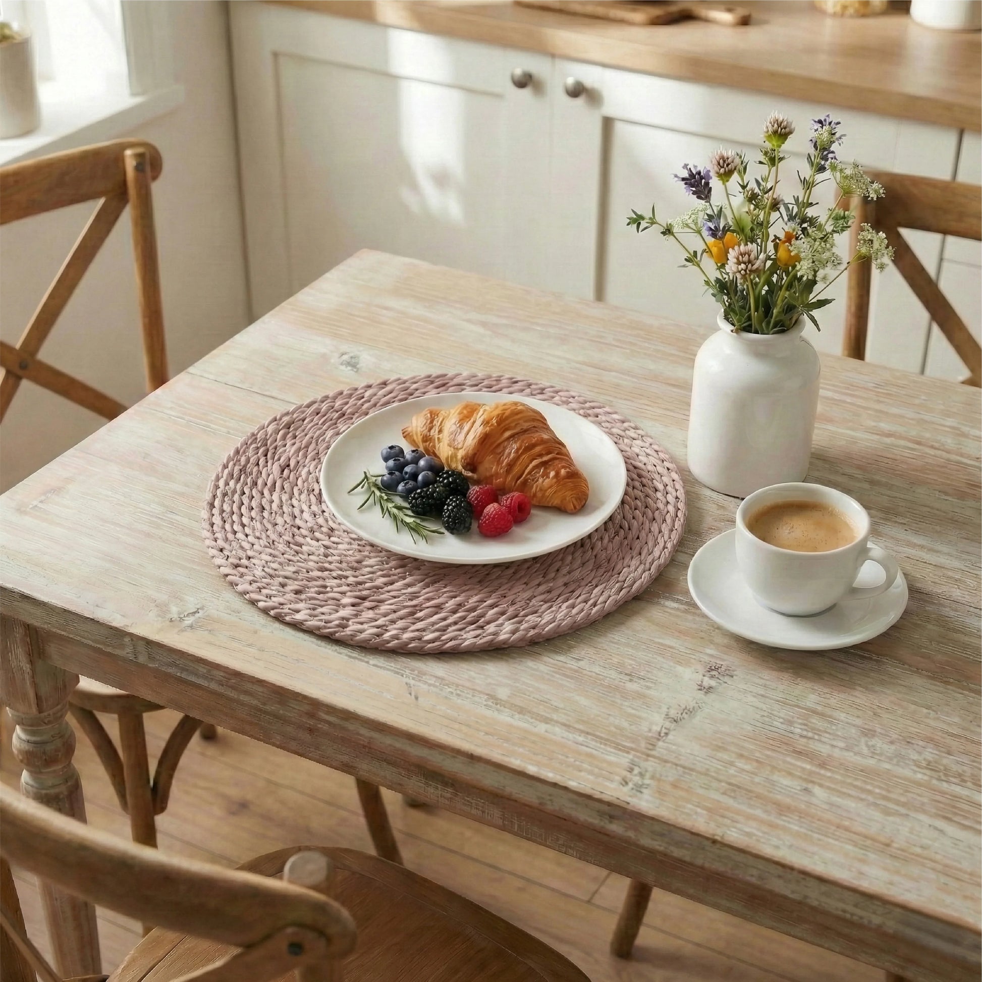 Pale pink halfa grass round placemat under a white plate with a croissant and berries, set on a distressed wood table with a coffee cup and wildflower vase.