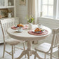 Round dining table set with croissants, coffee, and a vase of flowers in a bright room.