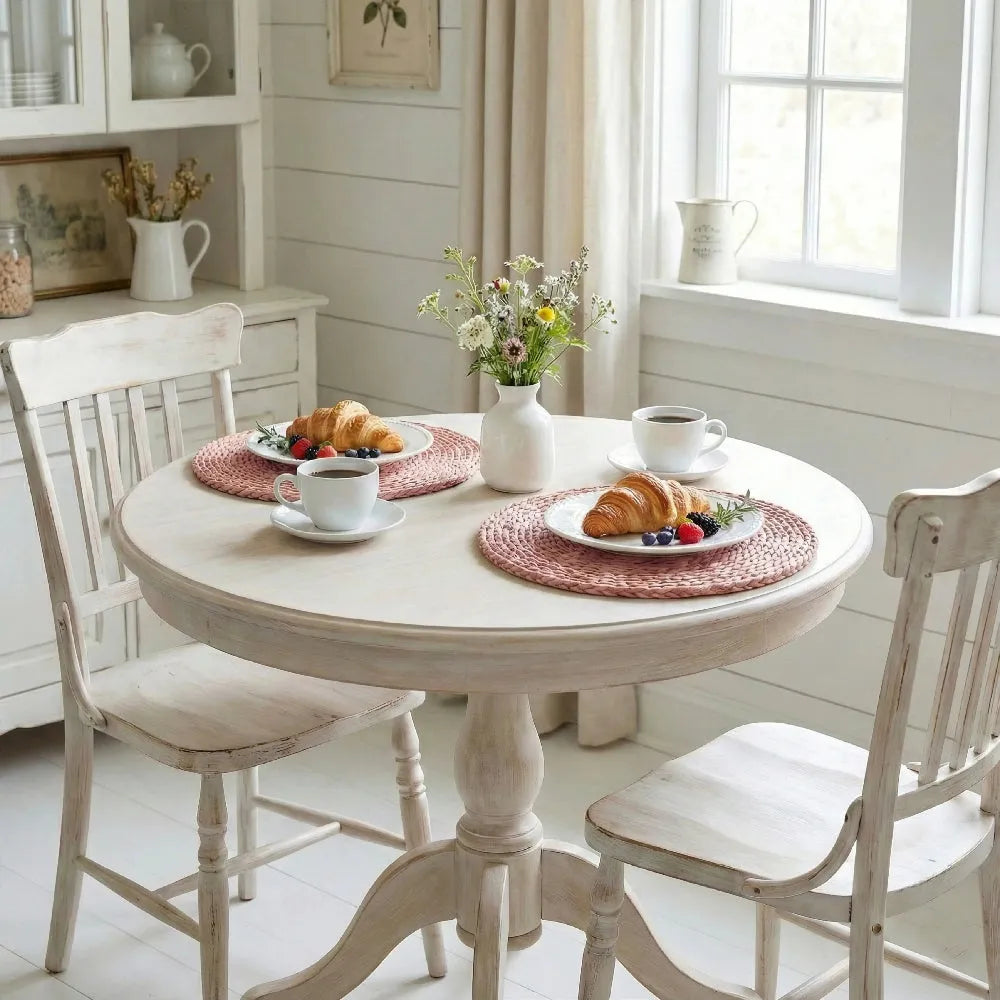 Round dining table set with croissants, coffee, and a vase of flowers in a bright room.
