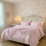 Pale pink ruffled bedding set with a matching duvet cover, cushions, and pillows on a white ornate bed frame.