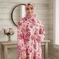 Woman wearing a pink floral dress and hijab in a room with wooden walls and a table with flowers.