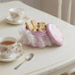 Pink cookie tin with lace detailing on a table with tea cups and cookies.