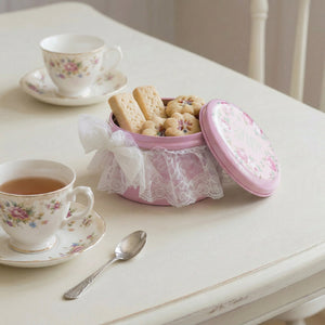 Pink cookie tin with lace detailing on a table with tea cups and cookies.