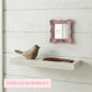 Small pink distressed mirror on a white plank wall above a floating shelf with a wooden bird and a brown comb.