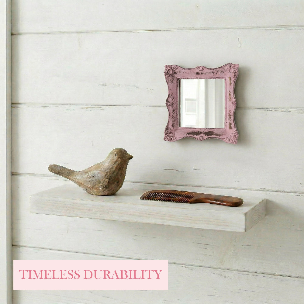 Small pink distressed mirror on a white plank wall above a floating shelf with a wooden bird and a brown comb.
