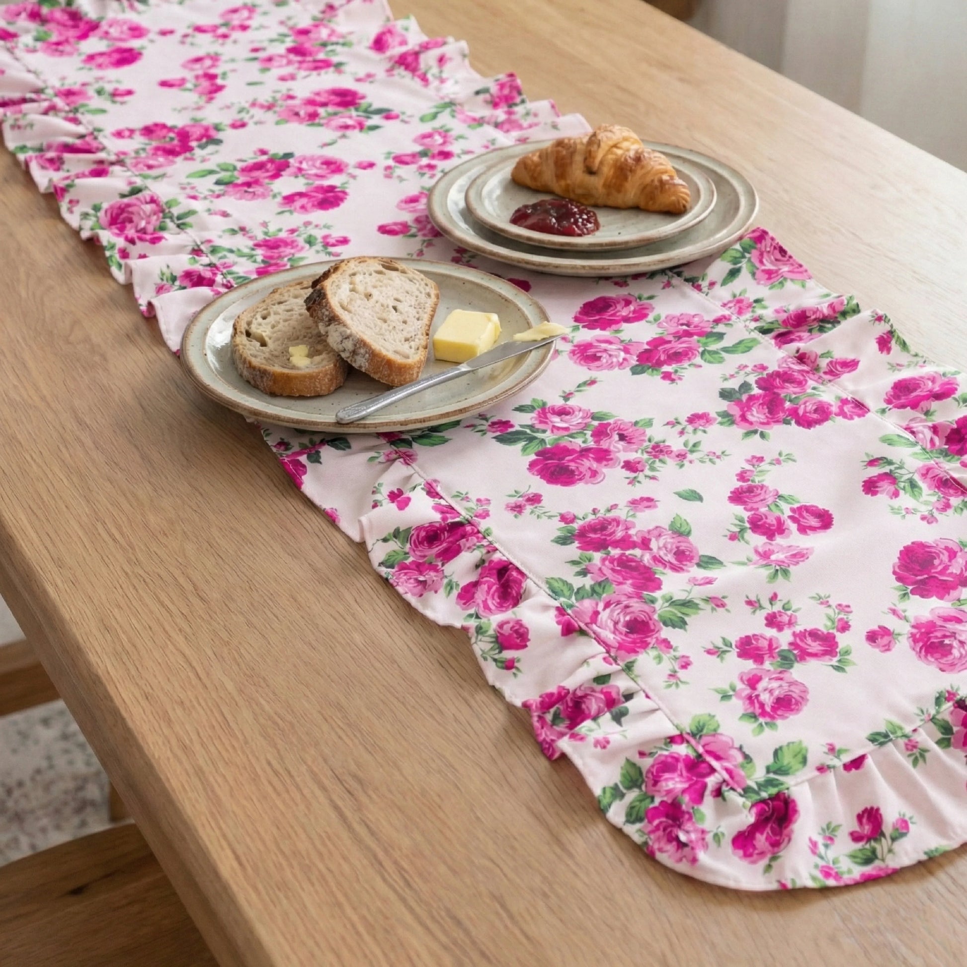Pink floral ruffled table runner on a wooden table with plates of bread, butter, a croissant, and jam.