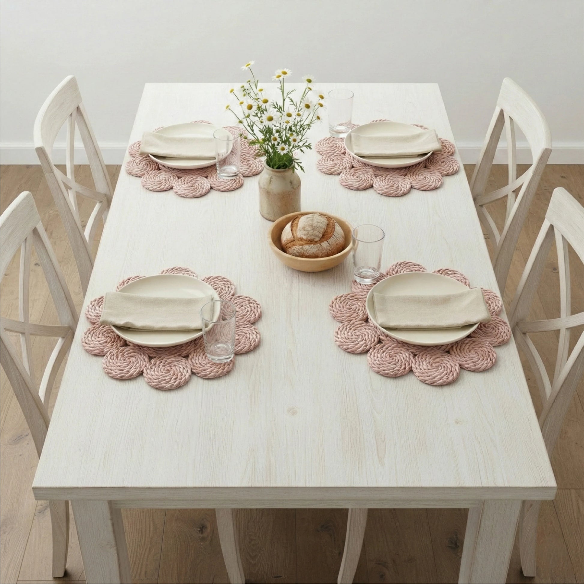 Four pink flower-shaped woven placemats with plates and napkins on an antique white table with a daisy centerpiece.